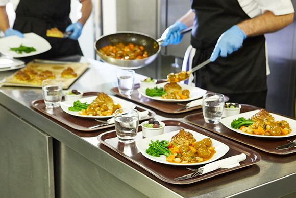 Ein Kantinenmitarbeiter verteilt Essensportionen auf Teller, die auf einem Kantinentablett liegen.