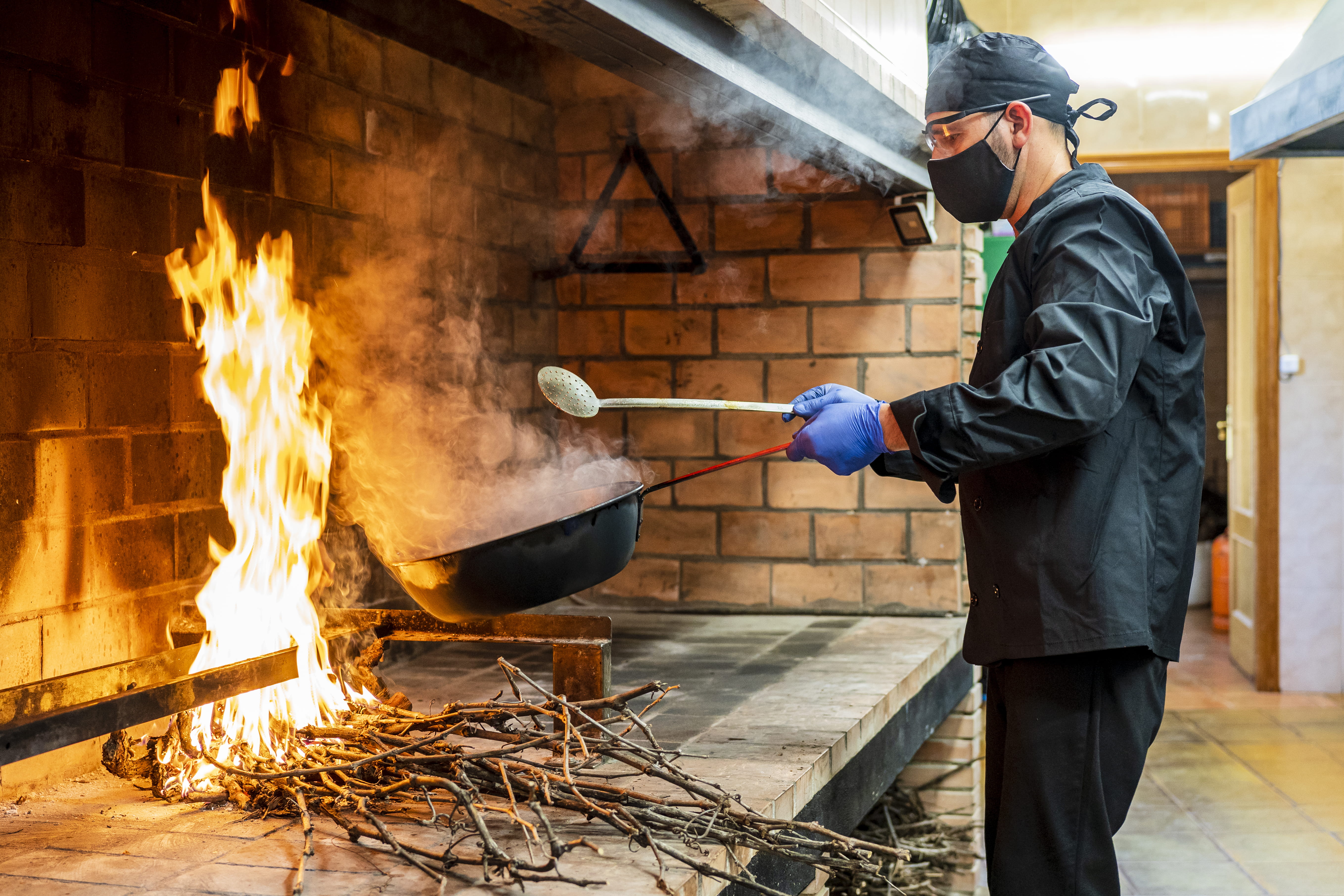 Professional chef wearing mask cooking paella outside kitchen on an open fire 